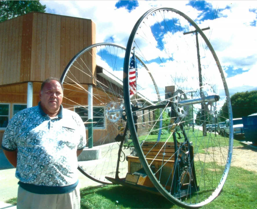 Man in blue patterned shirt standing next to a large bicycle wheel with blue sky and industrial building in background